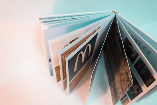 An open, colorful photo book with several pages displaying images, viewed from an angled side perspective. The pages appear to contain photographs and possibly text, with one page prominently featuring a logo that resembles an 'M'. The overall color scheme is light with a soft focus in parts of the image.