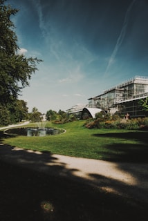 A large, modern greenhouse structure with glass panels stands amidst a lush garden setting. A small, tranquil pond with a water fountain is surrounded by well-manicured grass and flower beds. The sky is clear with streaks of clouds, and tall trees provide partial shade along the pathway.