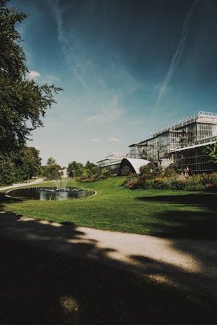 A large, modern greenhouse structure with glass panels stands amidst a lush garden setting. A small, tranquil pond with a water fountain is surrounded by well-manicured grass and flower beds. The sky is clear with streaks of clouds, and tall trees provide partial shade along the pathway.