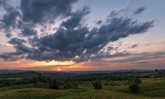 An expansive landscape photograph featuring rolling hills under a dramatic sky at sunset.