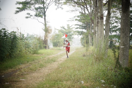 A person is running along a dirt path surrounded by lush green trees and grass. They are carrying a large red and white flag, and their outfit matches the flag. The pathway is lined with tall trees on the right and dense greenery on the left, creating a natural corridor.