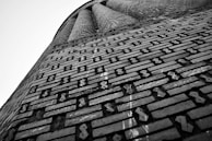 An architectural structure with intricate brickwork, featuring a geometric pattern. The bricks are arranged in a regular and repetitive design, with diagonal and vertical lines creating a visually striking texture. The perspective is angled upwards, giving a sense of height and grandeur.
