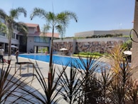 A swimming pool surrounded by a modern building with multiple stories, featuring large windows and a decorative wall. Palm trees and various plants are positioned around the pool, while people are casually walking or sitting near the water. The sky is clear and blue, adding a vibrant and sunny atmosphere.