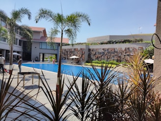 A swimming pool surrounded by a modern building with multiple stories, featuring large windows and a decorative wall. Palm trees and various plants are positioned around the pool, while people are casually walking or sitting near the water. The sky is clear and blue, adding a vibrant and sunny atmosphere.