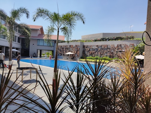 A swimming pool surrounded by a modern building with multiple stories, featuring large windows and a decorative wall. Palm trees and various plants are positioned around the pool, while people are casually walking or sitting near the water. The sky is clear and blue, adding a vibrant and sunny atmosphere.