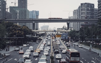 A busy urban scene features multiple lanes filled with cars and buses, with several overpasses crisscrossing overhead. Surrounding the road are tall buildings with modern architecture and construction cranes, indicating ongoing development. There are trees and some pedestrians visible along the sides.