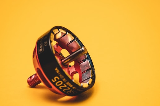 A component that appears to be a rotary device or motor part is positioned against a solid yellow background. The device is predominantly red and gold, displaying a shiny metal surface with circular openings around its circumference.