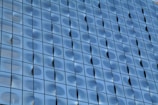 A detailed view of a modern glass building facade with a wavy, reflective surface. The glass panels are arranged in a grid pattern, creating a uniform yet dynamic appearance.