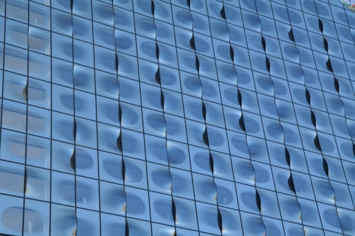 A detailed view of a modern glass building facade with a wavy, reflective surface. The glass panels are arranged in a grid pattern, creating a uniform yet dynamic appearance.