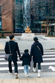 A family of three, consisting of two adults and a child, walk hand in hand across a zebra crossing. They are dressed in winter clothing, including coats and boots. In the background, there is a bronze statue of a seated figure on a pedestal, set against a modern glass building with reflective surfaces. Leafless trees and neatly trimmed shrubs are also visible.
