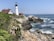 A picturesque coastal scene featuring a tall, white lighthouse with a black top, surrounded by buildings with red roofs. The lighthouse stands on rocky cliffs covered with greenery, overlooking the ocean. Waves crash against the rocks under a clear blue sky.