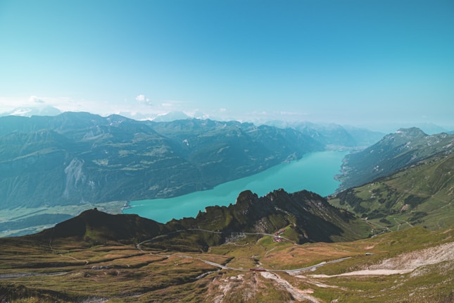 A stunning panoramic view of a mountainous landscape with a serene turquoise lake at its center. The scene features rugged mountain ranges with lush greenery in the foreground and distant snowy peaks under a clear blue sky.