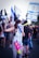 A group of people are gathered, holding Israeli flags. One person in the foreground has covered their face partially with a flag and is wearing striped pants, carrying a colorful toy water gun. The crowd seems engaged in a protest or demonstration as they wave flags and banners.