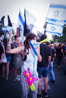A group of people are gathered, holding Israeli flags. One person in the foreground has covered their face partially with a flag and is wearing striped pants, carrying a colorful toy water gun. The crowd seems engaged in a protest or demonstration as they wave flags and banners.