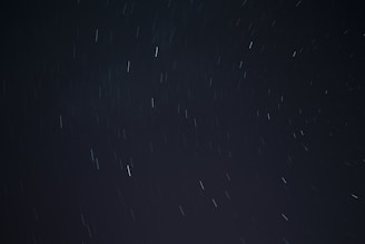 A long exposure photograph of the night sky, displaying streaks of light from stars or celestial bodies moving due to the rotation of the Earth. The background is dark with scattered lines of light across the frame.