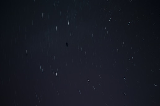A long exposure photograph of the night sky, displaying streaks of light from stars or celestial bodies moving due to the rotation of the Earth. The background is dark with scattered lines of light across the frame.