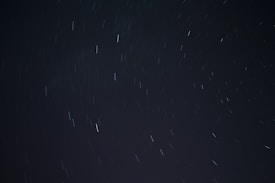 A long exposure photograph of the night sky, displaying streaks of light from stars or celestial bodies moving due to the rotation of the Earth. The background is dark with scattered lines of light across the frame.