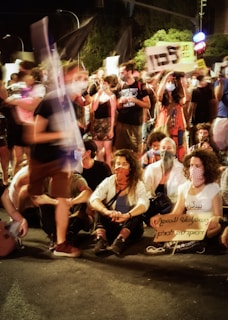 A large group of people gathered in a public space during what appears to be a protest or demonstration. Many individuals are standing while others are sitting on the ground. Most of them are wearing face masks, and some are holding signs and banners with text. The atmosphere suggests a collective gathering with visible expressions of activism.
