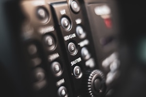 Close-up of a camera control panel featuring various buttons and dials with labels such as ISO/GAIN and SHUTTER. The focus is shallow, blurring parts of the panel and creating a soft, artistic effect. The lighting appears dim and warm, emphasizing the details of the buttons and surfaces.