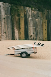 A trailer is parked on a sandy area, carrying several surfboards secured with orange straps. In the background, a large concrete wall with patches of greenery partially covers a hill.