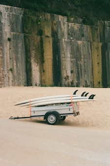 A trailer is parked on a sandy area, carrying several surfboards secured with orange straps. In the background, a large concrete wall with patches of greenery partially covers a hill.