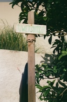 A wooden post is wrapped with rope just below a small rectangular sign reading 'Wetsuits' in green letters. The post is set against a backdrop of lush green foliage and a beige stucco wall. Nearby, part of a dark fabric, likely a wetsuit, is hanging.