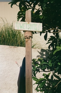 A wooden post is wrapped with rope just below a small rectangular sign reading 'Wetsuits' in green letters. The post is set against a backdrop of lush green foliage and a beige stucco wall. Nearby, part of a dark fabric, likely a wetsuit, is hanging.