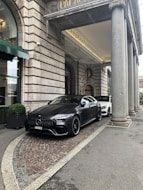 A luxury car is parked in front of an ornate, historic building with large stone columns. The vehicle is sleek and modern, painted in a matte black shade. The entrance to the building features a green awning and decorative stonework.