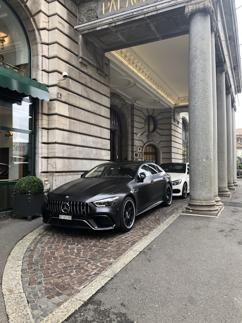 A luxury car is parked in front of an ornate, historic building with large stone columns. The vehicle is sleek and modern, painted in a matte black shade. The entrance to the building features a green awning and decorative stonework.