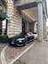 A luxury car is parked in front of an ornate, historic building with large stone columns. The vehicle is sleek and modern, painted in a matte black shade. The entrance to the building features a green awning and decorative stonework.