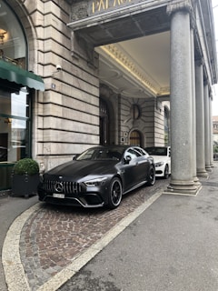 A luxury car is parked in front of an ornate, historic building with large stone columns. The vehicle is sleek and modern, painted in a matte black shade. The entrance to the building features a green awning and decorative stonework.