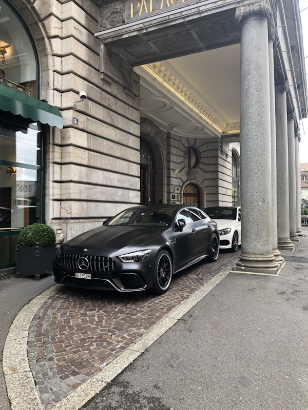A luxury car is parked in front of an ornate, historic building with large stone columns. The vehicle is sleek and modern, painted in a matte black shade. The entrance to the building features a green awning and decorative stonework.