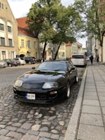 A black sports car is parked on a cobblestone street in a European city. The surroundings include several historic buildings with light-colored plaster and red tile roofs. A large tree provides shade on the sidewalk, and people are walking along the street. Other parked cars line the street, contributing to the urban atmosphere.