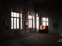 A dimly lit room with large windows allowing soft natural light to pour in, casting shadows on the floor. A vintage upright piano sits against the wall, partially illuminated. The walls are decorated with framed pictures, and the decor suggests an ambient, possibly artistic or historical space.