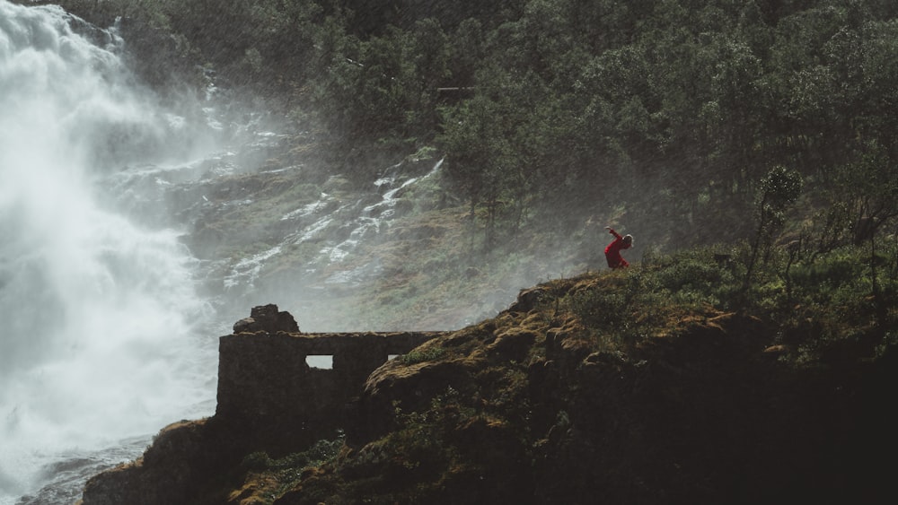 person in red jacket standing on rock near water falls during daytime