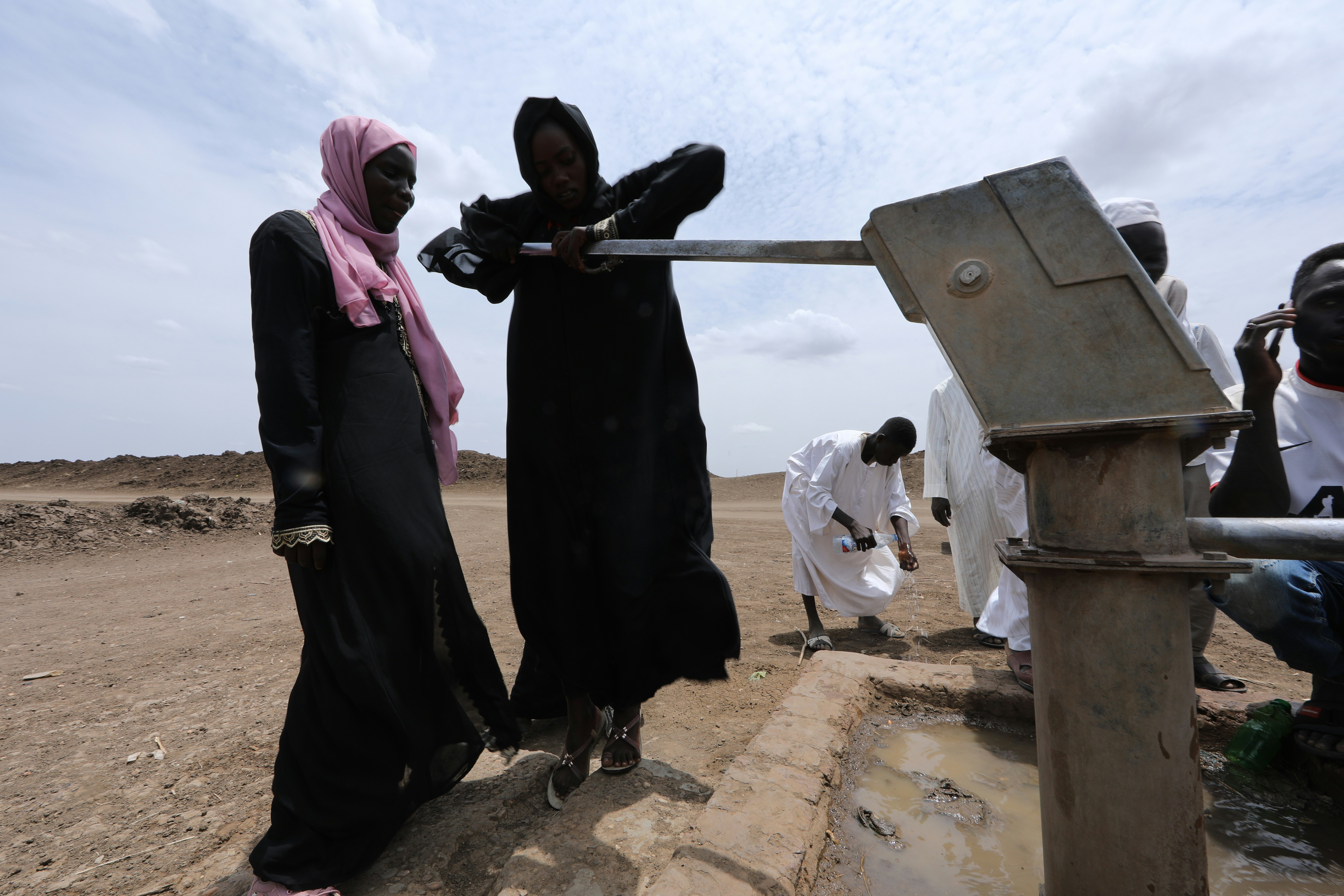 African woman at a clean water borehole pump, dusty village setting, bright midday sun