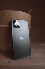 A modern smartphone with a sleek design is leaning against a wooden bucket. The phone's back is visible, showcasing its metallic finish, multiple camera lenses, and a prominent logo. The setting appears dimly lit, creating highlights on the reflective surfaces.