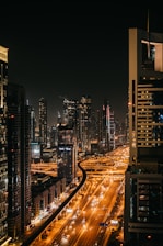A cityscape at night with a network of illuminated roads and towering skyscrapers. The buildings are lit up with various lights, and the streets are bustling with vehicles, creating a dynamic urban scene.