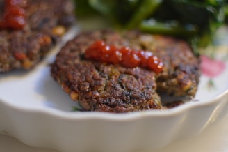 A close-up of two browned patties topped with a dollop of red sauce on a white plate, accompanied by green leafy vegetables in the background.