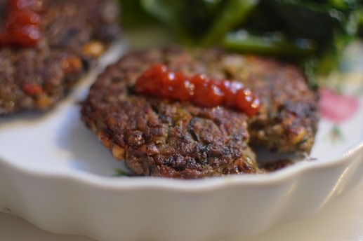 A close-up of two browned patties topped with a dollop of red sauce on a white plate, accompanied by green leafy vegetables in the background.