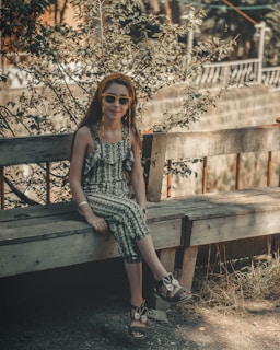 A child wearing sunglasses and a patterned jumpsuit sits on a wooden bench. The setting is outdoors, surrounded by leafy plants and muted colors. The child has a headband and is smiling slightly, suggesting a relaxed and casual moment on a sunny day.