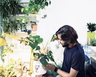 A person with medium-length hair is tending to potted plants indoors, pouring water from a small watering can. The space is filled with a variety of green plants, both hanging and placed on shelves. The scene is well-lit, with a yellow desk lamp adding to the bright ambiance.