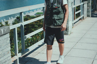 A person is standing on a paved walkway next to a railing overlooking a body of water. The individual is wearing a green utility vest over a white shirt and dark shorts with white sneakers. The background includes greenery and a view of a coastline.