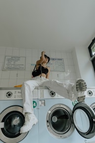 A person dressed in a black top and white pants is sitting on top of a row of washing machines in a laundromat. Their posture is relaxed, with one foot planted on the machine and the other hanging loosely. The laundry area is clean with tiled walls, and there's a window letting in natural light.