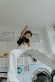 A person sits on top of a washing machine in a laundry room. They are wearing white pants, a black crop top, and a patterned headscarf. The room has white tiled walls with instructions posted above the washing machines.