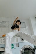 A person sits on top of a washing machine in a laundry room. They are wearing white pants, a black crop top, and a patterned headscarf. The room has white tiled walls with instructions posted above the washing machines.