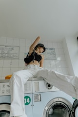 A person sits on top of a washing machine in a laundry room. They are wearing white pants, a black crop top, and a patterned headscarf. The room has white tiled walls with instructions posted above the washing machines.