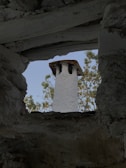 A stone chimney with a rustic appearance is viewed through an opening framed by rough, textured stone walls. The backdrop features green foliage against a clear blue sky.