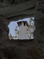 A stone chimney with a rustic appearance is viewed through an opening framed by rough, textured stone walls. The backdrop features green foliage against a clear blue sky.