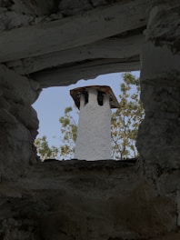 A stone chimney with a rustic appearance is viewed through an opening framed by rough, textured stone walls. The backdrop features green foliage against a clear blue sky.